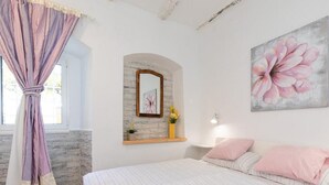 Un dormitorio con cama, ventana con cortinas, espejo montado en la pared, un cuadro de flores y una pequeña estantería con un florero.