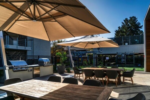 An outdoor patio with a wooden table and chairs, a barbecue grill, and large umbrellas providing shade.