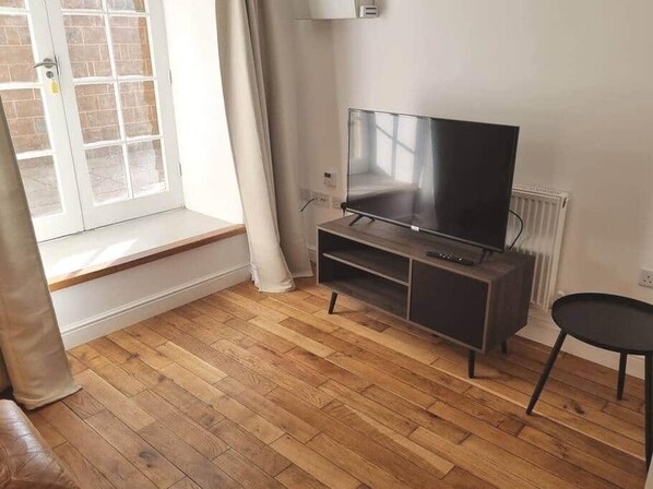 Une pièce avec un plancher en bois, un téléviseur posé sur un meuble, un radiateur et une fenêtre avec des rideaux.