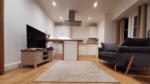 Una sala de estar moderna con televisor de pantalla plana, un sofá oscuro y una cocina con gabinetes blancos y una isla central.
