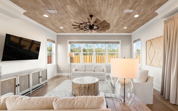 A spacious living room with a modern ceiling fan, a large flat-screen TV mounted on the wall, and a large window with a view of the outdoors.