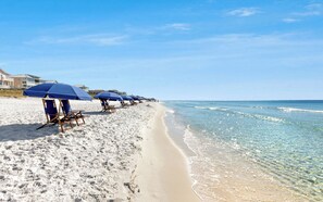 A beach with rows of blue beach chairs and umbrellas, a sandy shoreline, and clear blue water.