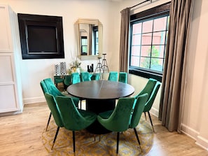 Uma área de jantar com uma mesa redonda, cadeiras estofadas em verde, um tapete estampado e uma janela com cortinas.