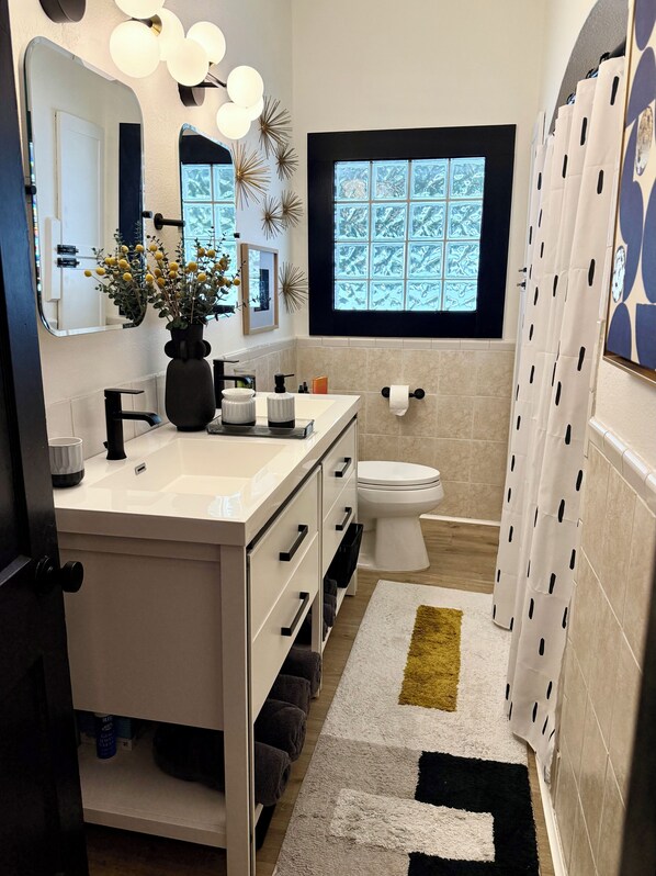 A modern bathroom with a white vanity, a toilet, a window with a view, and a shower curtain with a pattern.