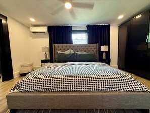 A bedroom with a large bed, a headboard, a nightstand with a lamp, a window with curtains, and an air conditioning unit on the wall.