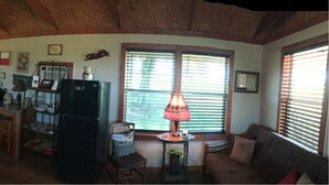 A cozy living room with a wooden ceiling, a dining area with a table and chairs, a kitchen with a refrigerator and stove, and a window with blinds. - Country Cabin  10 miles from Round Top (Burton)