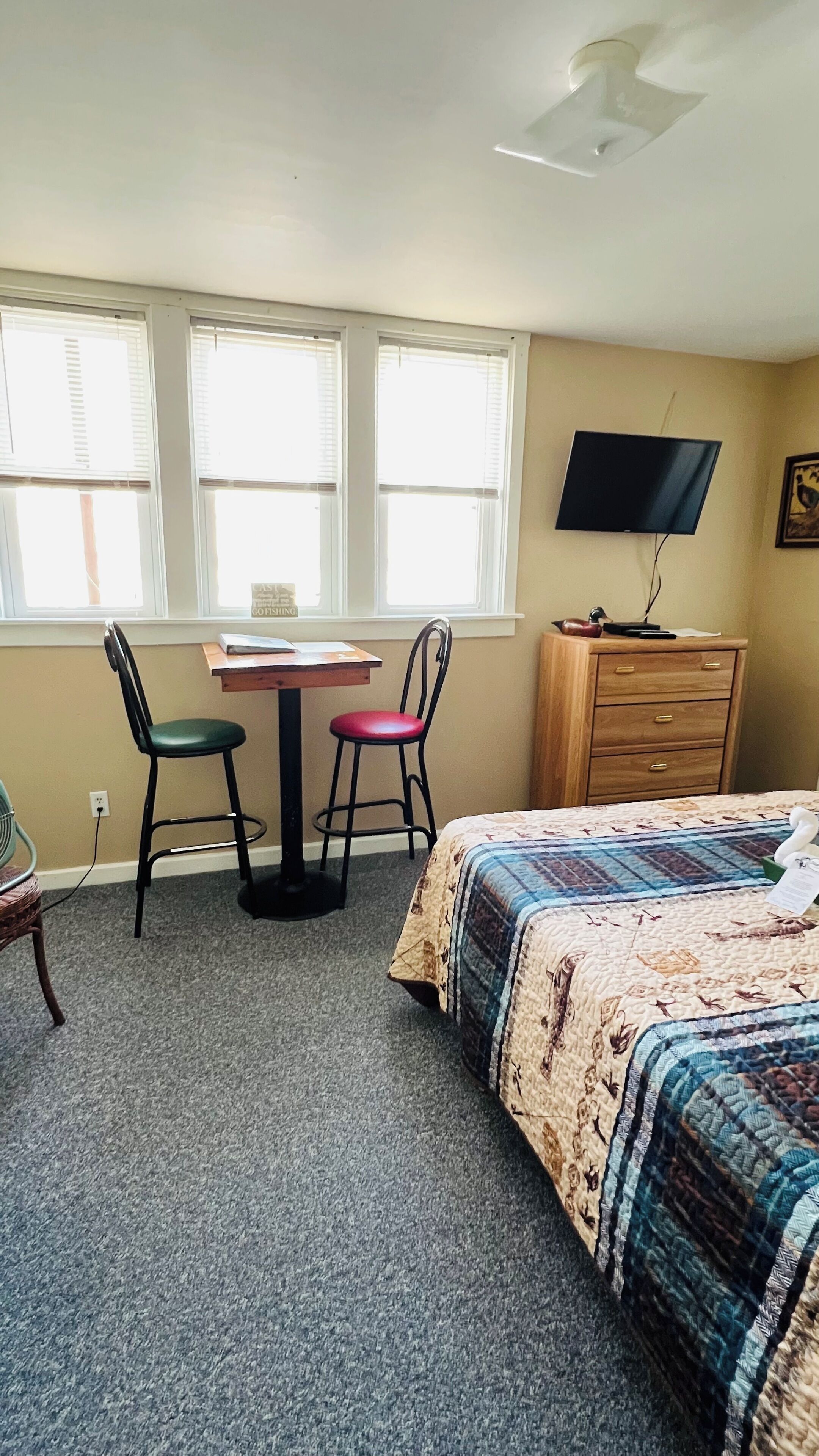 A bedroom with a bed, a desk, two chairs, a dresser, and a TV on the wall.