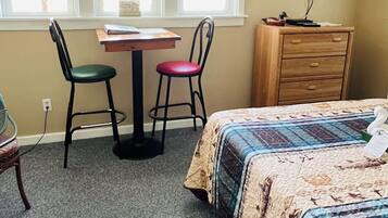 A bedroom with a bed, a desk, two chairs, a dresser, and a TV on the wall.