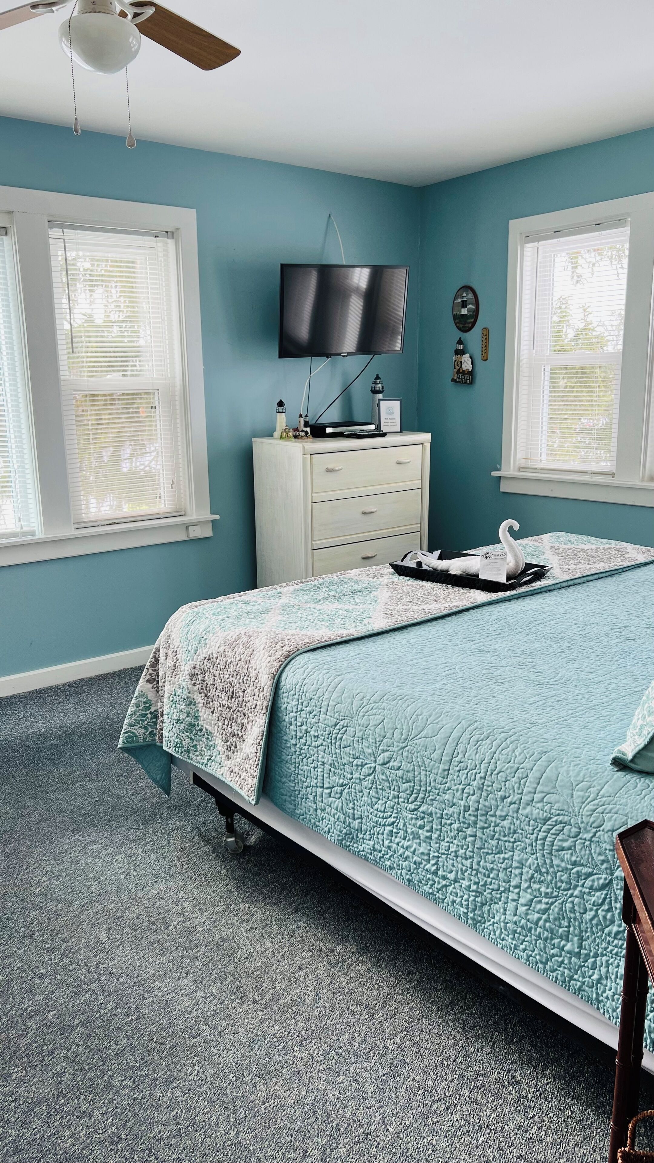 Une chambre à coucher comprenant un lit, une commode, un téléviseur et deux fenêtres équipées de stores.