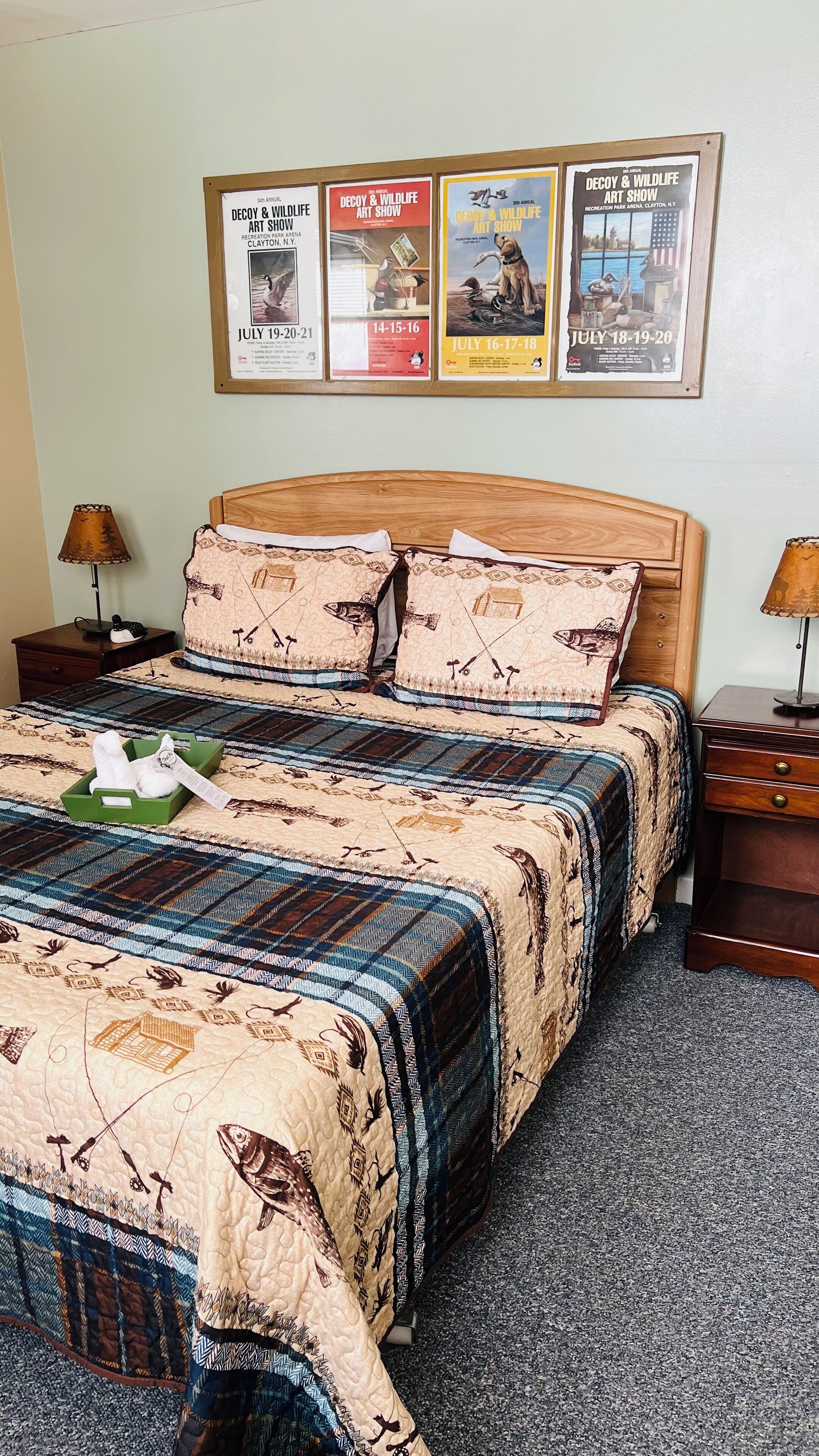 A neatly made bed with a quilt featuring fish and fishing hooks, flanked by two nightstands with lamps, and a wall displaying three framed posters of the Decoy & Wildlife Art Show.