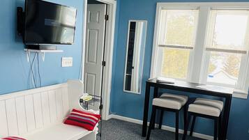A room with a blue wall, a white couch with red cushions, a TV mounted on the wall, a door, a window with blinds, and a small table with two chairs.