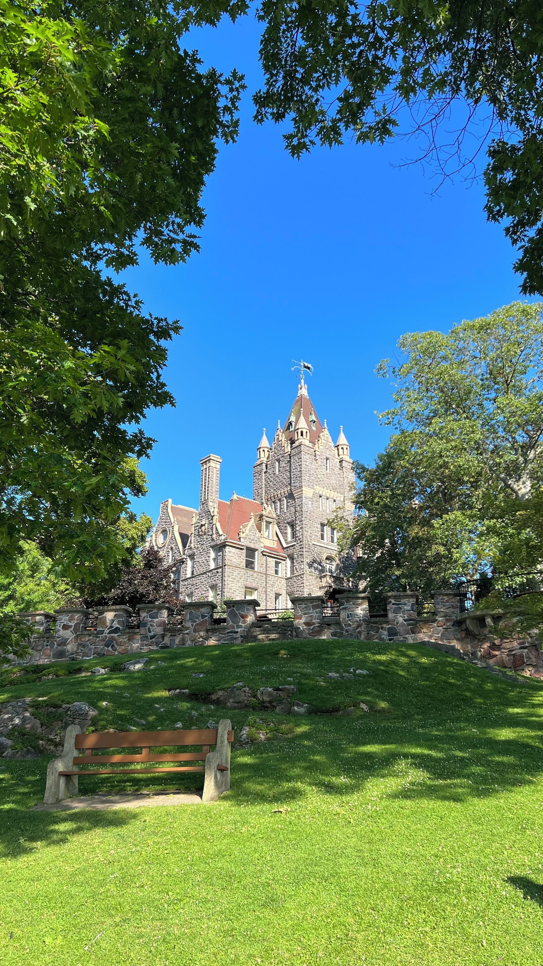 A historic stone building with a steep roof and multiple towers, surrounded by lush green trees and a grassy area with a bench.