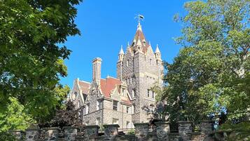 A historic stone building with a steep roof and multiple towers, surrounded by lush green trees and a grassy area with a bench.