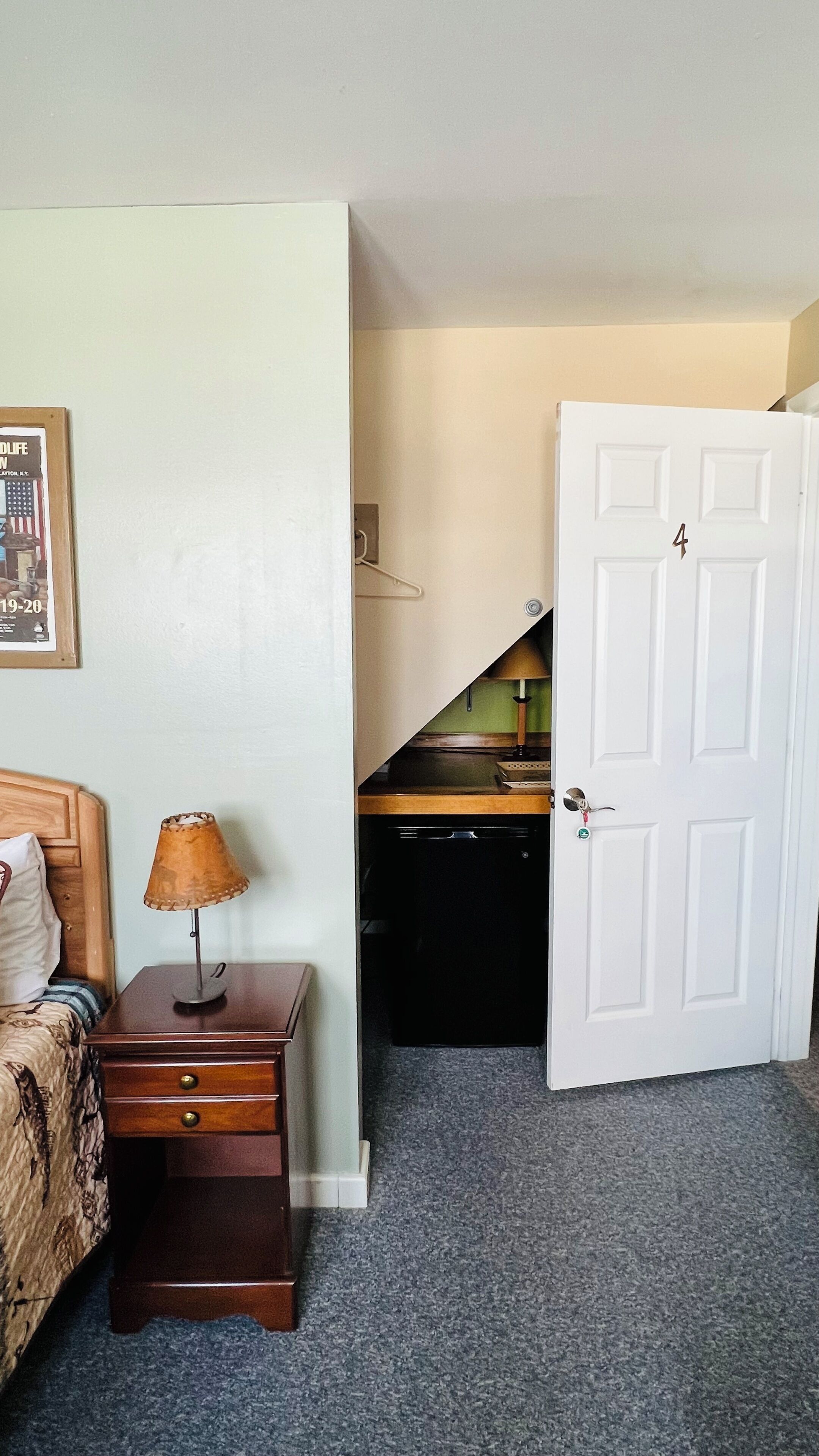 A bedroom with a bed, a nightstand with a lamp, a door, and a desk with a chair.