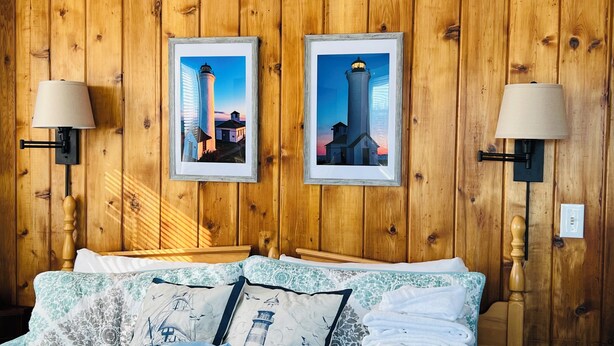 A wooden-paneled room with two framed pictures of lighthouses, a bed with floral bedding, and wall-mounted lamps. - Chateau Country Inn  (Clayton)