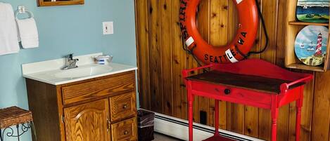 A bathroom with wooden walls, a wooden vanity, a red table, a life preserver, and a shelf with decorative items.