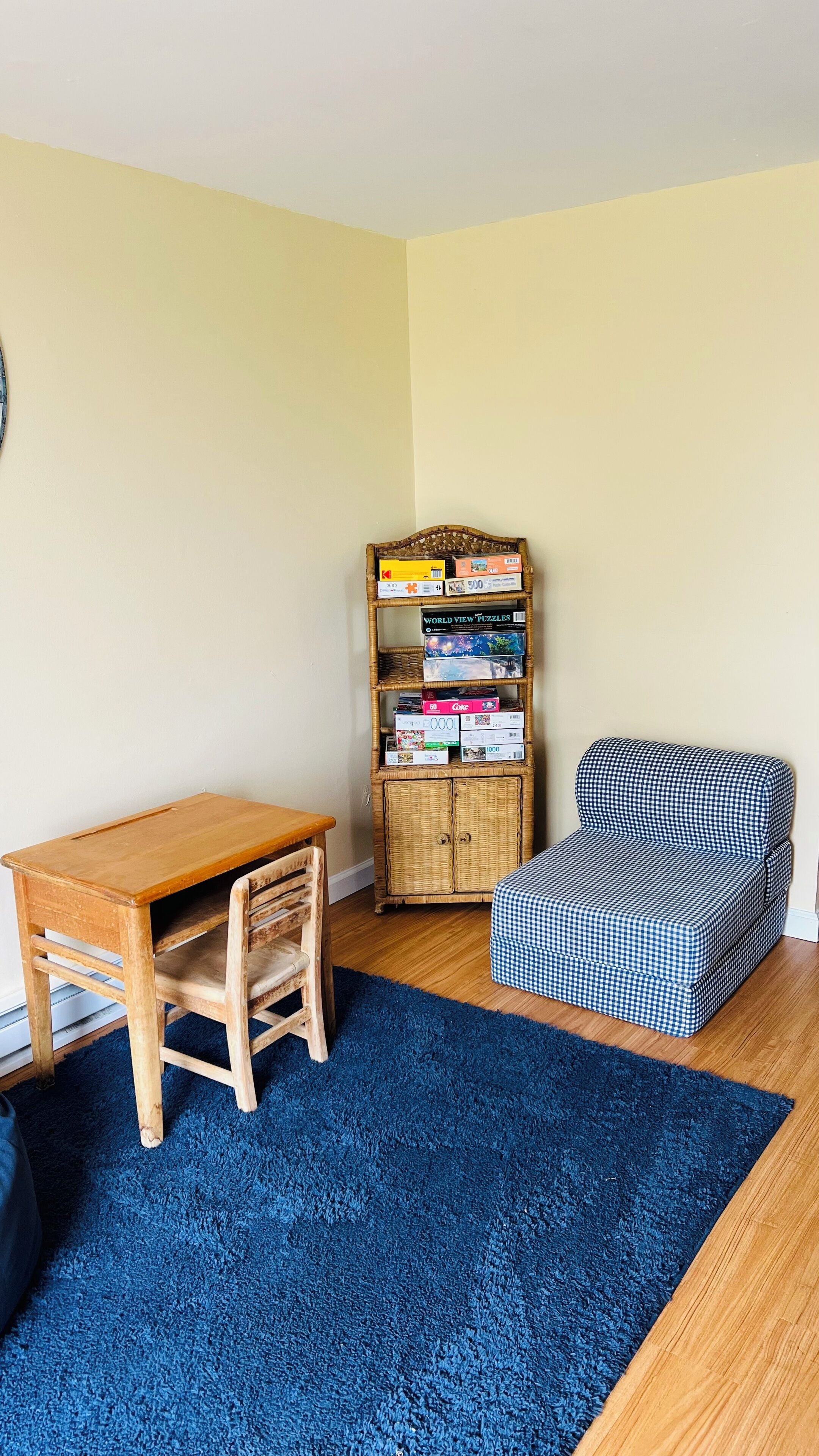 Een kamer met een houten tafel, een blauw vloerkleed, een rieten boekenkast en een geruit voetenbankje.