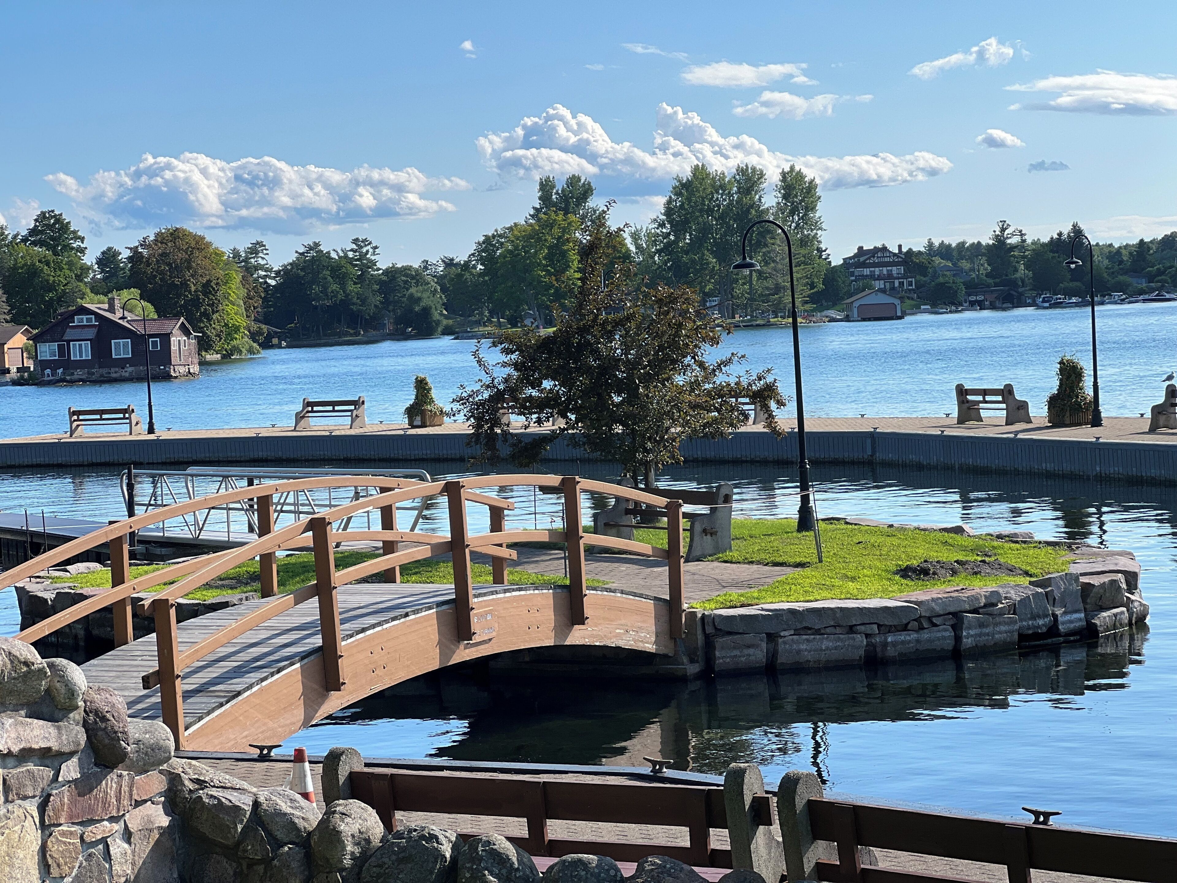A wooden bridge over a calm body of water, surrounded by greenery and a clear sky.