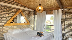 A bedroom with a brick wall, a wooden ceiling, a bed with white bedding, a hanging light fixture, and a window with curtains. - Villa Flecheiras  (Trairi)