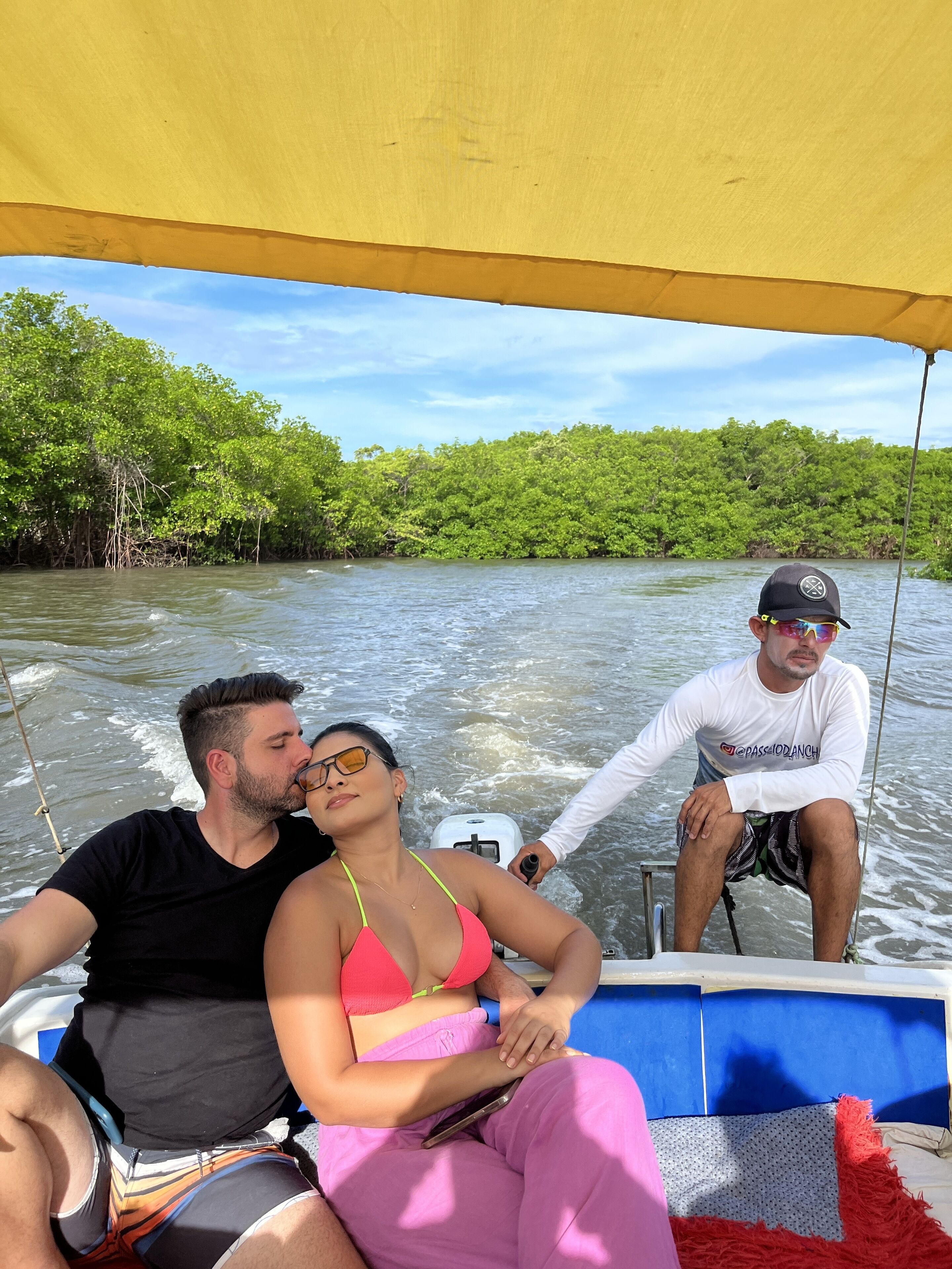 A boat with a yellow canopy, a man in a white shirt and black cap, and a woman in a pink bikini and pink pants.