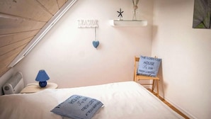 A bedroom with a sloped ceiling, a bed with white bedding, a blue pillow, a small table with a lamp, a wall-mounted shelf with decorative items, and a chair with a sign. - FH „Kajüte“81m², 4 Pers. im Marinapark Scharmützelsee, Sauna, Whirlpool, Kamin (Wendisch Rietz Siedlung)