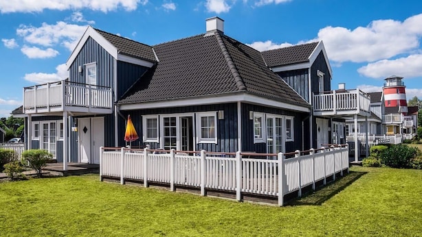A two-story house with a balcony, a white fence, and a red and white lighthouse in the background. - FH „Kajüte“81m², 4 Pers. im Marinapark Scharmützelsee, Sauna, Whirlpool, Kamin (Wendisch Rietz Siedlung)