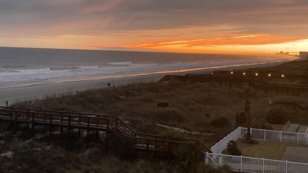 A coastal view with a wooden boardwalk leading to a beach, overlooking the ocean and a city skyline in the distance. - Ocean front at the chateau (North Myrtle beach)
