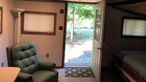 A room with a wooden ceiling, a green cushioned chair, a small table, and a door leading outside. - Little White Cabin by the river, fishing trails, kayaking, near State Park lake  (Manns Choice)