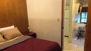 Ein Schlafzimmer mit einem Bett, einem Nachttisch und einer Lampe. Durch eine geöffnete Tür ist ein Badezimmer zu sehen.