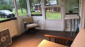 Ein Zimmer mit einer hölzernen Bank, einem Stuhl und einem Ventilator. Eine Tür führt nach draußen, mit Blick auf ein Haus und ein Auto.