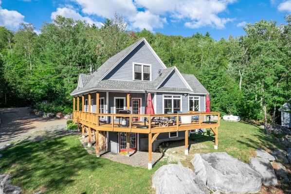 A two-story house with a deck, surrounded by a forest.