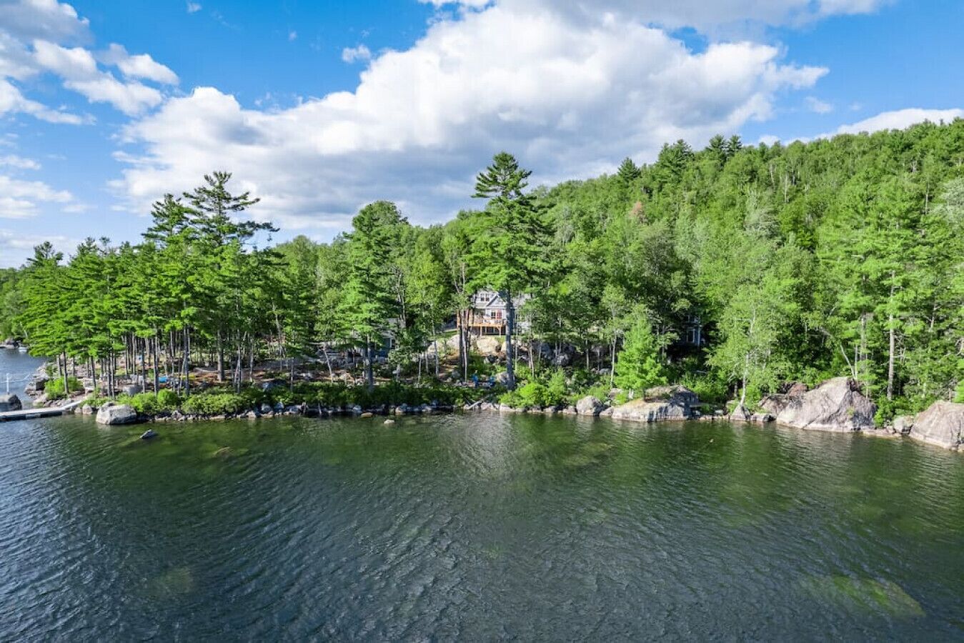 A lakeside property with a large house surrounded by dense forest, a dock with boats, and a clear sky.