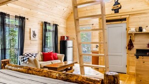 En mysig stuga med träinredning, en säng av trä, en soffa med röda kuddar, ett litet bord och en stege som leder upp till en dörr.