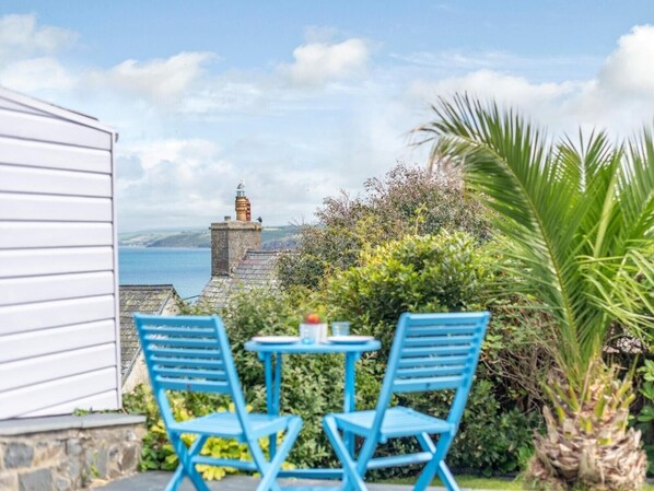 A patio with two blue chairs and a small table, overlooking a coastal view with a stone building and a chimney.