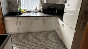 A kitchen with white cabinets, a tiled floor, a window, and a ceiling light fixture. - Small, cozy semi-detached house on a large plot (Heide)