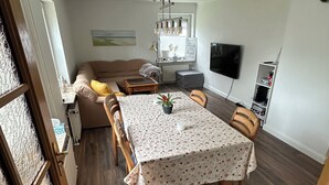 A dining area with a table set for four, a sofa, a TV, and a kitchen in the background. - Small, cozy semi-detached house on a large plot (Heide)