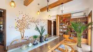 A dining area with a long wooden table, chairs, and hanging pendant lights. There is a bookshelf filled with books and decorative items. - wuginggu Hostel (Hangzhou)