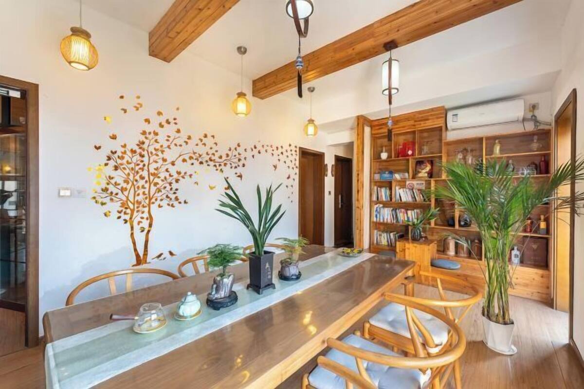 A dining area with a long wooden table, chairs, and hanging pendant lights. There is a bookshelf filled with books and decorative items.