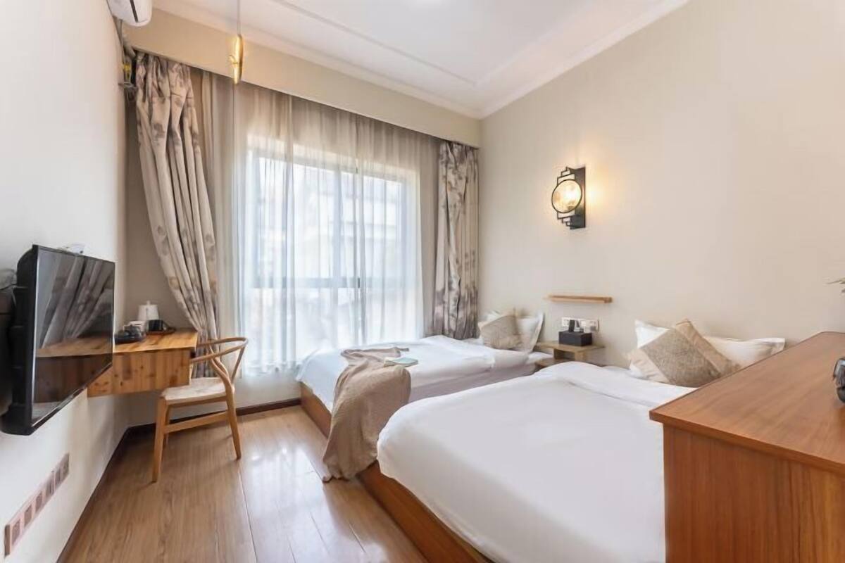 A hotel room with two beds, a desk, a chair, a TV, and a wall-mounted light fixture.