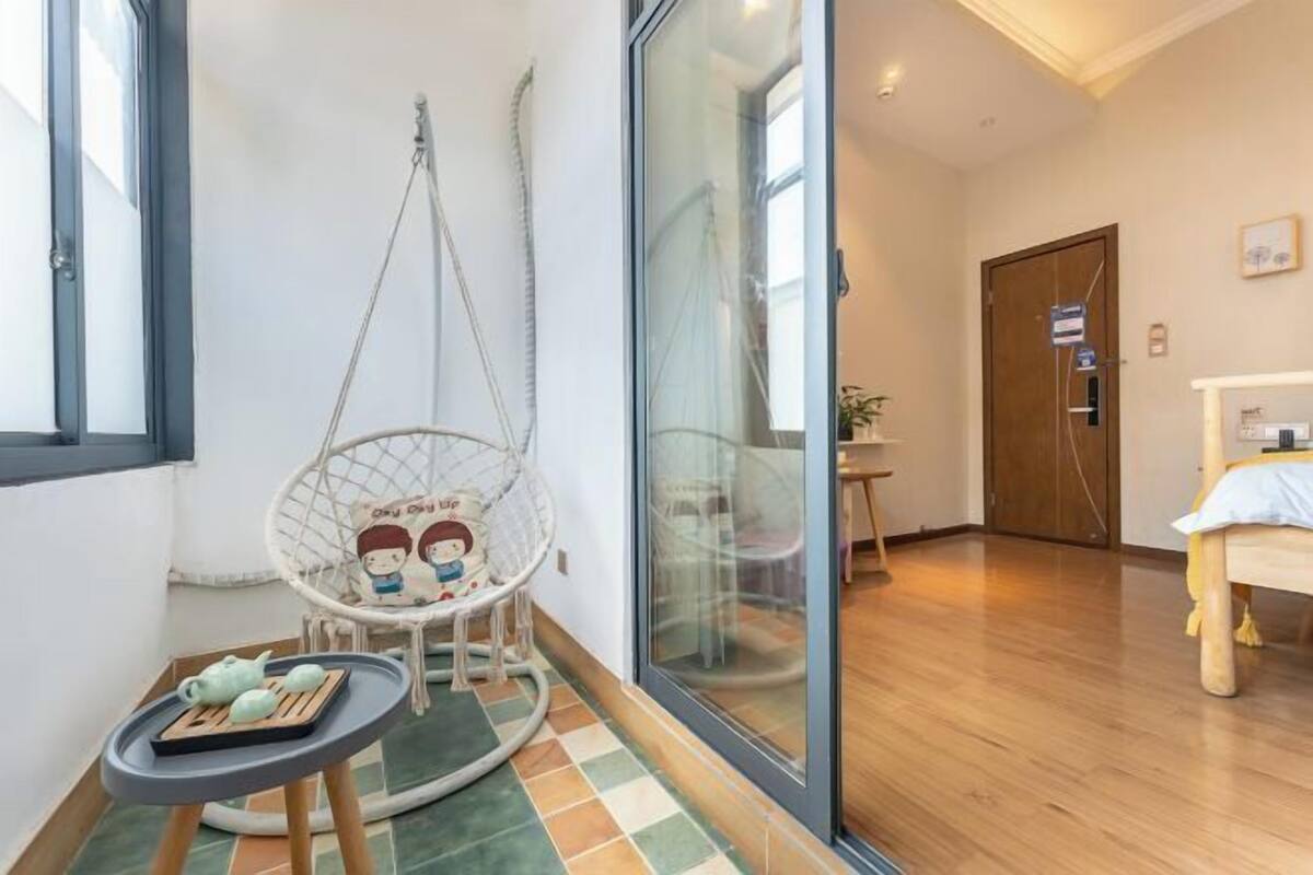 A room with a large sliding glass door, a wooden floor, a bed with a white and yellow bedspread, a small table with a teapot and cups, and a hanging chair with a cartoon character design.