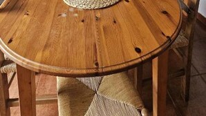 A round wooden dining table with chairs, a painting above it, and a kitchen area with cabinets and appliances in the background. - Apartment in the countryside (Coaña)