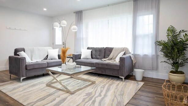 Una sala de estar moderna con dos sofás grises, una mesa de centro de vidrio y un ventanal con cortinas translúcidas.