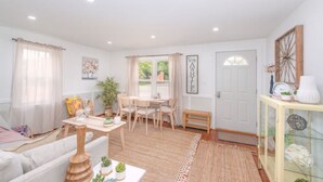 A cozy living room with a sofa, a dining table with chairs, a coffee table, and a small cabinet. There is a window with curtains, a door, and a wall-mounted decoration.