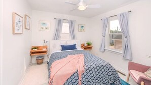 A bedroom with a bed, a ceiling fan, a window with curtains, a bedside table with a lamp, and a small plant.
