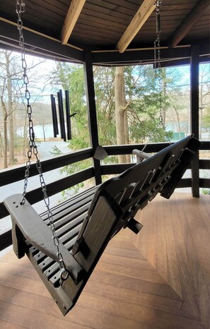 A wooden swing suspended by chains in a covered outdoor area with a view of trees and a body of water.
