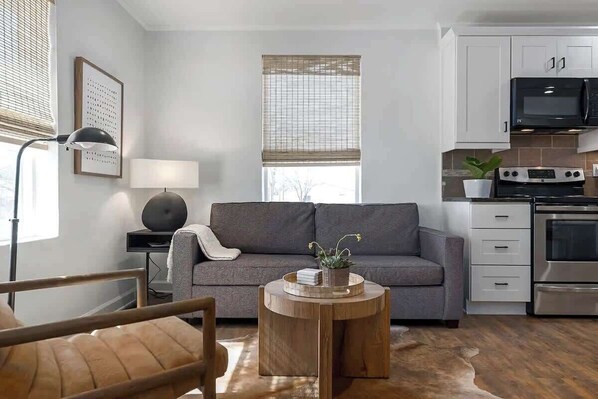 Ein modernes Wohnzimmer mit grauem Sofa, einem runden Couchtisch, einem Essbereich mit Tisch und Stühlen, einer Küche mit weißen Schränken und Edelstahlgeräten sowie großen Fenstern mit Jalousien.