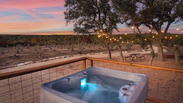 Un jacuzzi en una terraza con vistas al atardecer, luces colgantes y una mesa de picnic.