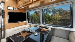 Interior de una autocaravana bien iluminada, con un televisor montado, una mesa decorada con estrellas y ventanas amplias que dan a un paisaje de árboles.