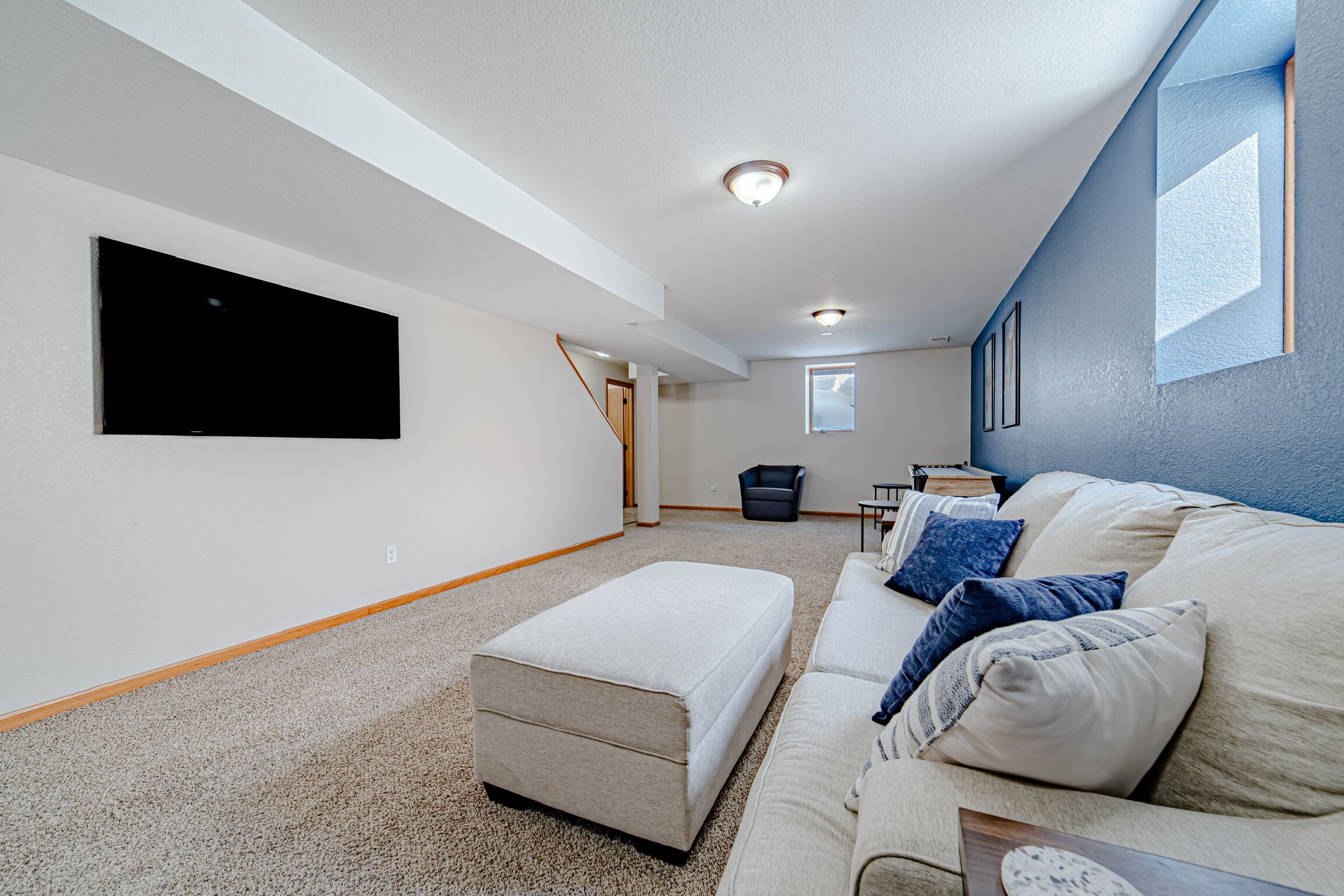 Ein geräumiges Wohnzimmer mit einem großen Flachbildfernseher, einem beigen Sofa mit blauen Kissen, einem weißen Hocker und einem kleinen Tisch mit Büchern.