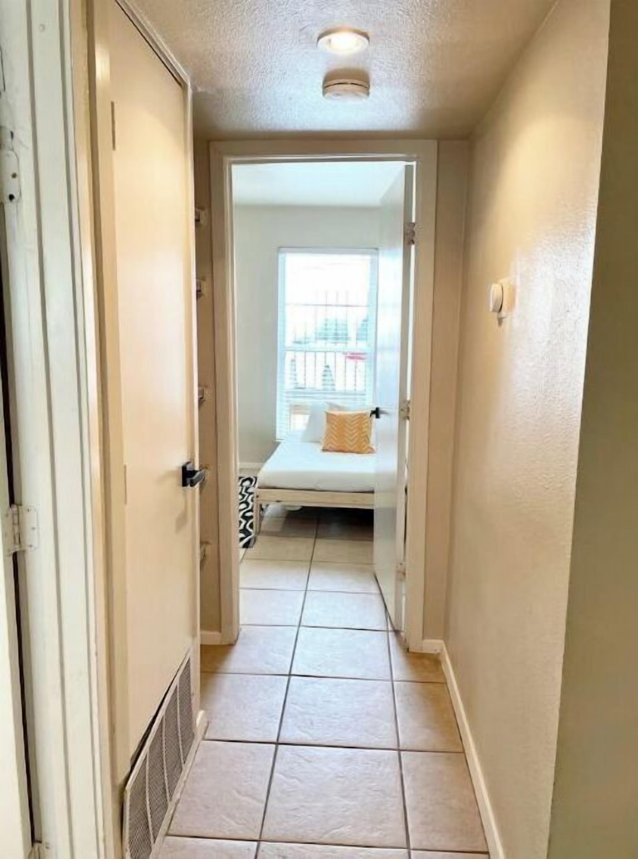A narrow hallway with a bed in the background, a window with a view of buildings, and a door on the left.
